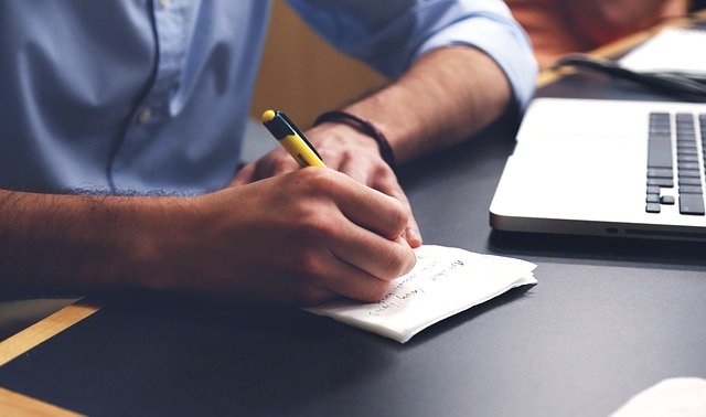 Mężczyzna w niebieskiej koszuli, który pisze na kartce papieru. Obok znajduje się laptop Fot. pixabay