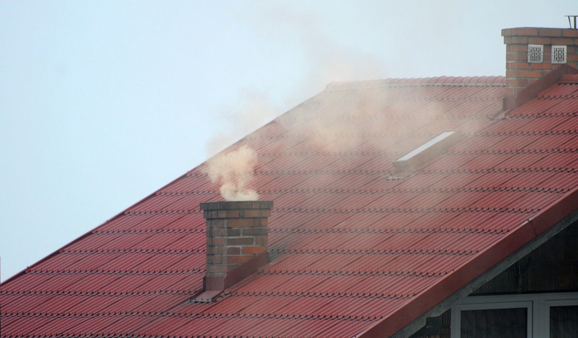 Zdjęcie przedstawia czerwony dach na budynku. Na dachu znajduje się komin, z którego wydobywa się dym. Zdjęcie: Pixabay