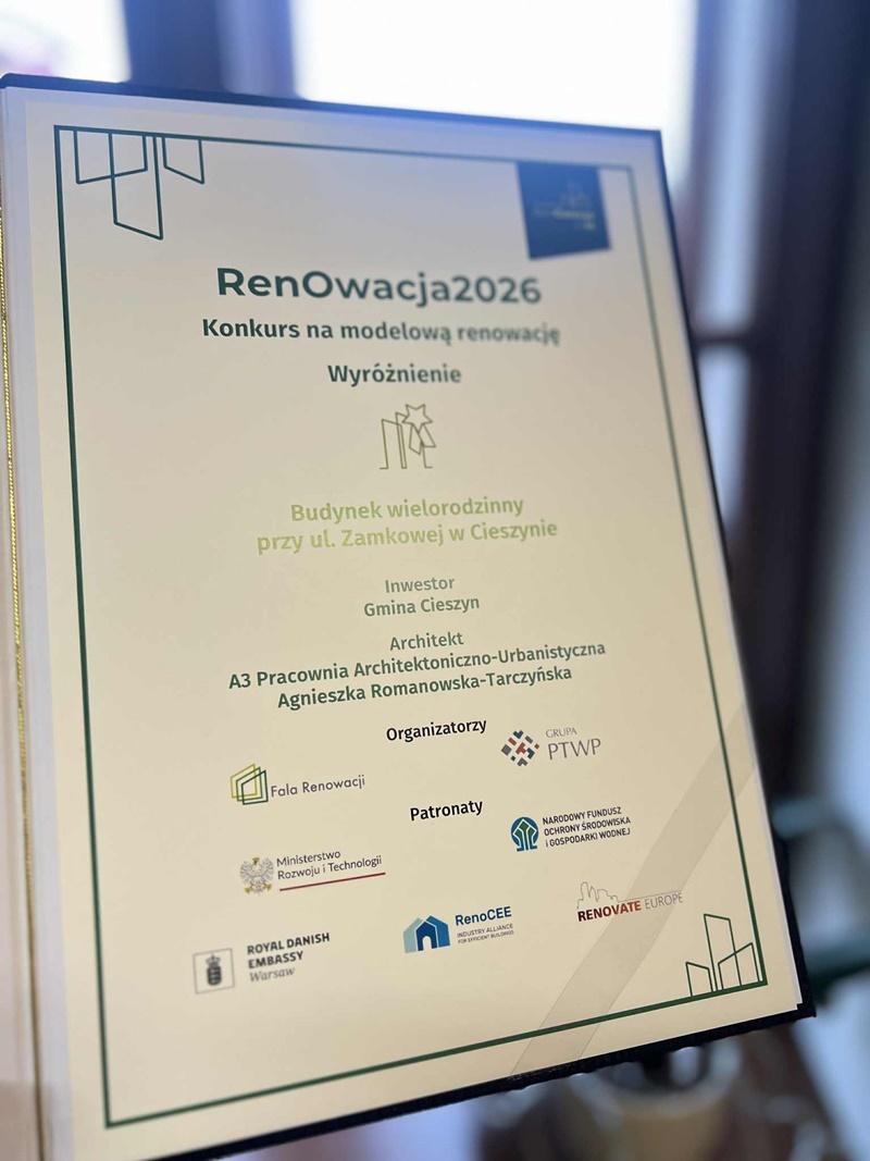 Zdjęcie przedstawia dyplom w jasnej ramce, ustawiony pionowo, na tle rozmytego wnętrza. Na dyplomie widoczny jest tytuł „RenOwacja2026” oraz informacja o konkursie na modelową renowację. Dokument przyznaje wyróżnienie dla budynku wielorodzinnego przy ul. 