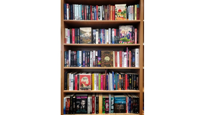 Drewniany regał z kilkoma półkami wypełnionymi książkami. Na półkach stoją różnorodne tytuły: powieści, reportaże, książki popularnonaukowe i dla dzieci, o kolorowych okładkach, fot. Biblioteka Miejska FB