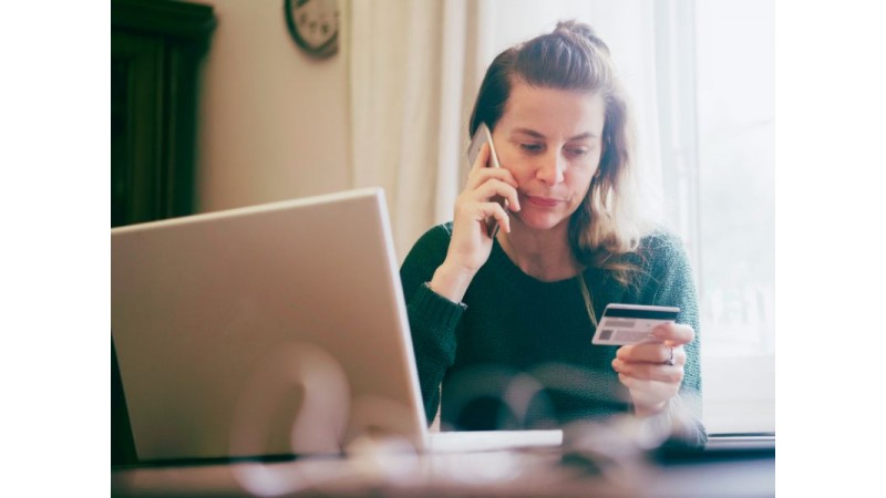 Kobieta w zielonym swetrze siedzi przy stoliku, rozmawiając przez telefon, trzymając w drugiej dłoni kartę bankomatową. Przed nią leży otwarty laptop. W tle widać okno z delikatnym światłem i zegar, tło jest rozmyte. fot. canva