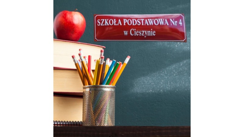 Na ławce stoją książki z czerwonym jabłkiem i kubek z kolorowymi ołówkami, w tle zielona tablica z napisem „Szkoła Podstawowa Nr 4 w Cieszynie”.
